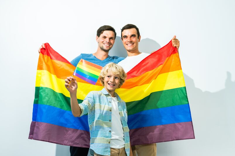Two men smile and hold a Pride flag behind their son, who happily looks on while holding  a smaller flag.