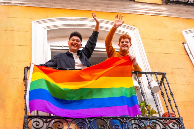 Two men smile and wave from a balcony while holding a Pride flag.