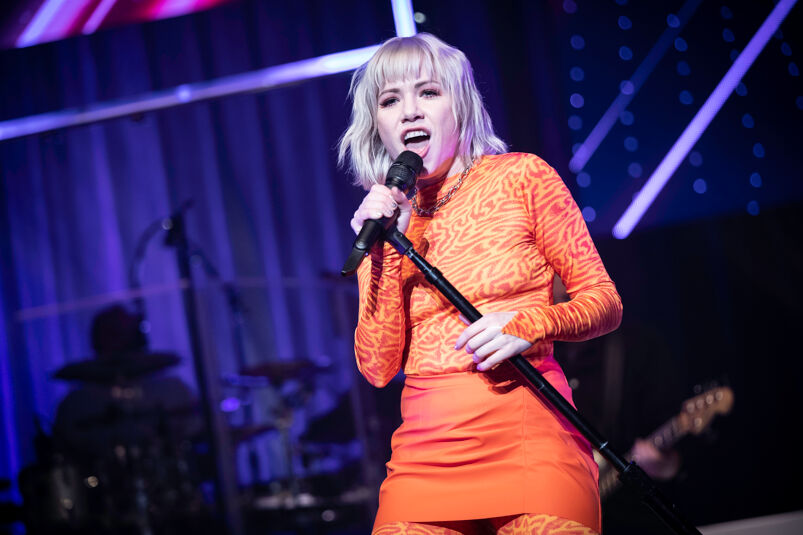 Carly Rae Jepsen singing on stage in a striped orange shirt and bright orange skirt. 
