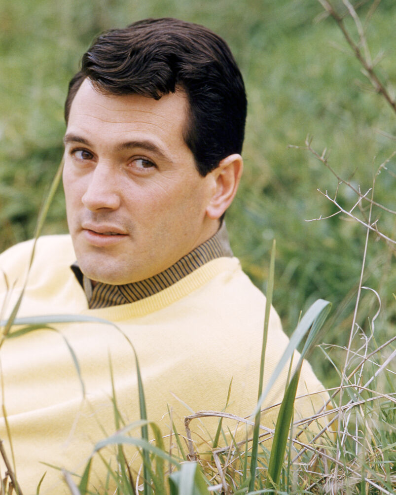 Rock Hudson, wearing a yellow v-neck jumper as he sits among grass in a publicity portrait, circa 1965.