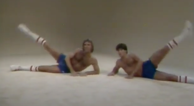 Two shirtless Chippendales wearing blue booty shorts in a workout video.