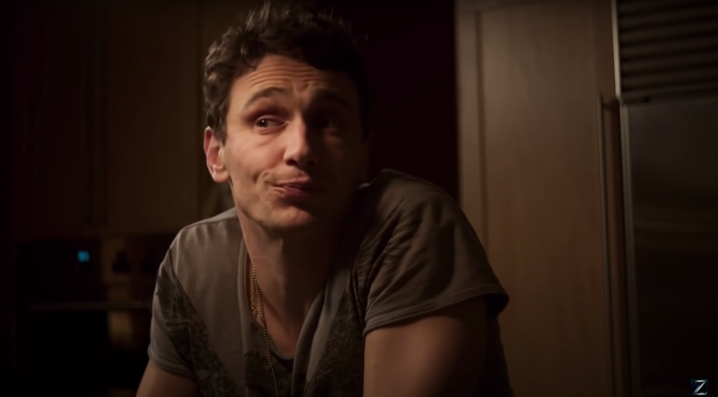 James Franco smirks sassily, wearing a gray colored tee, in moody lighting as a gay porn producer in 2016's 'King Cobra.'