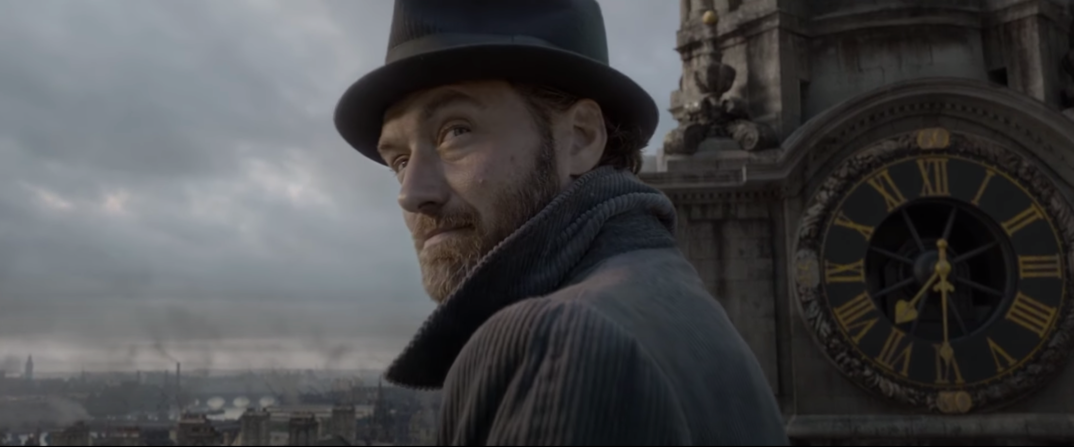 Jude Law, wearing an overcoat and top hat, looks over his shoulder in front of a clock tower as Albus Dumbledore in a scene from 'Fantastic Beasts.'