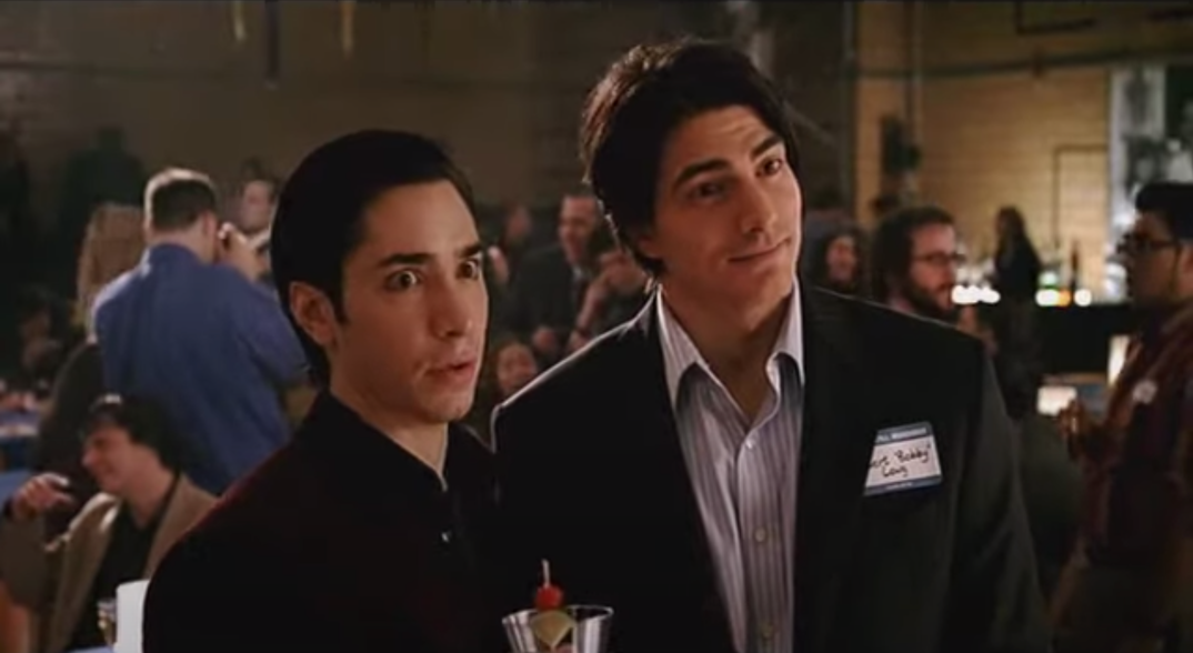 Justin Long, holding a martini, and dressed in a black dress shirt, stands along Brandon Routh, wearing a name tag and suit jacket, at a high school reunion in a scene from 'Zack and Miri Make a Porno.'
