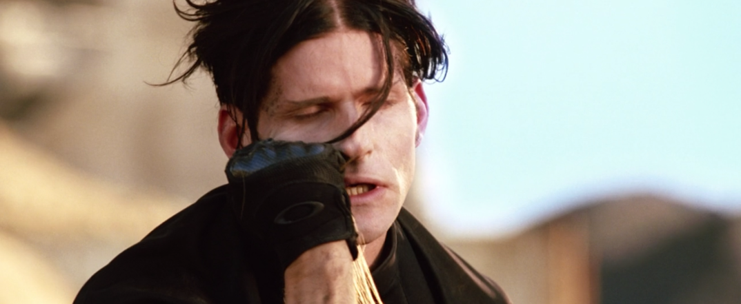 Crispin Glover, with messy black hair and a black glove, smells a strand of hair after falling off a bike in 'Charlie's Angels: Full Throttle.'
