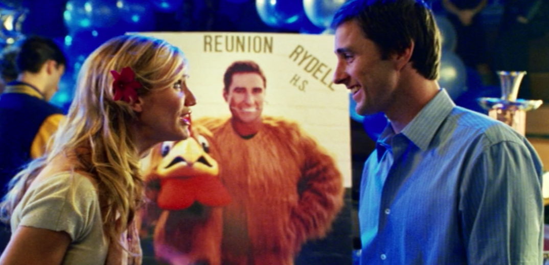 Cameron Diaz looks happily at Luke Wilson in front of a photo of him dressed as a rooster at a high school reunion.