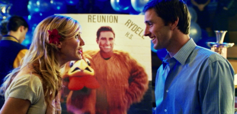 Cameron Diaz looks happily at Luke Wilson in front of a photo of him dressed as a rooster at a high school reunion.