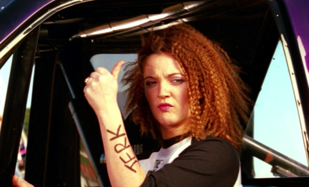 Drew Barrymore, with frizzy red hair and dark lipstick, gives a thumbs up in the driver's seat of a monster truck. The word "JERK" is written on her arm in Sharpie.