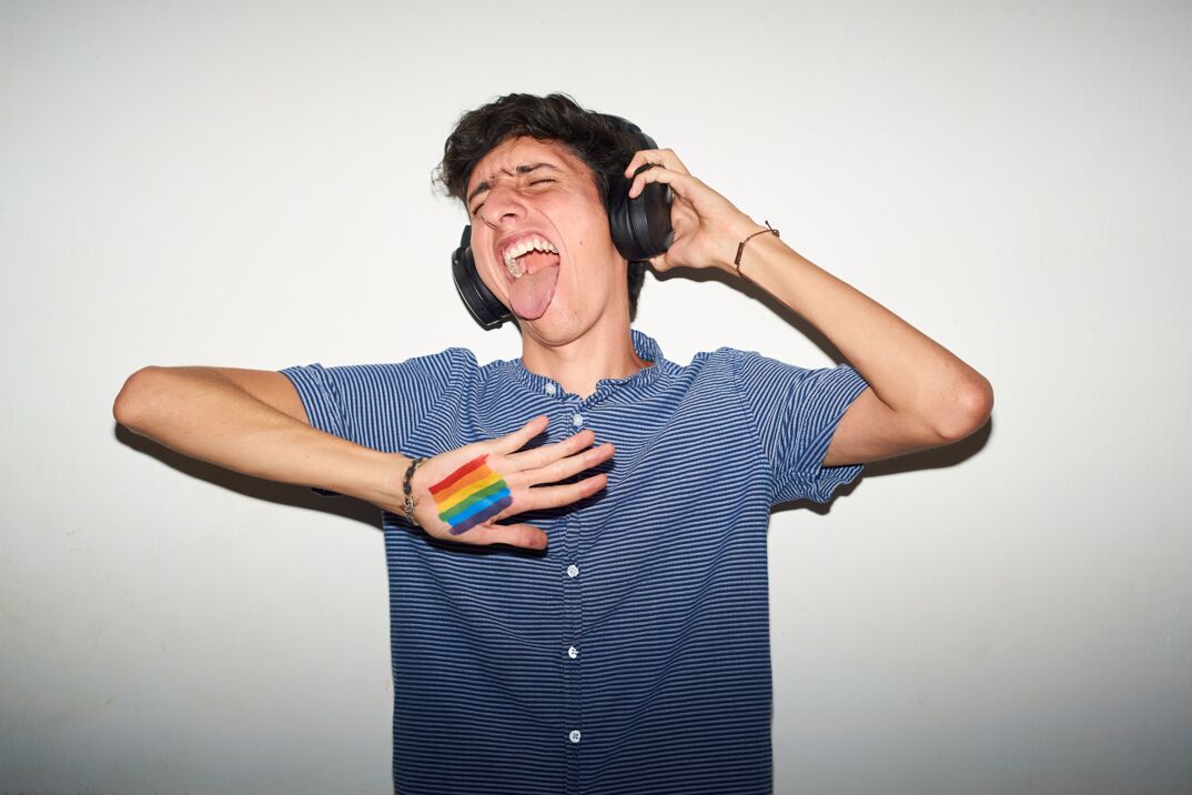 Cheerful white boy in his early 20s wearing black headphones and screaming while listening to music, with a rainbow flag superimposed on his open palm. 
