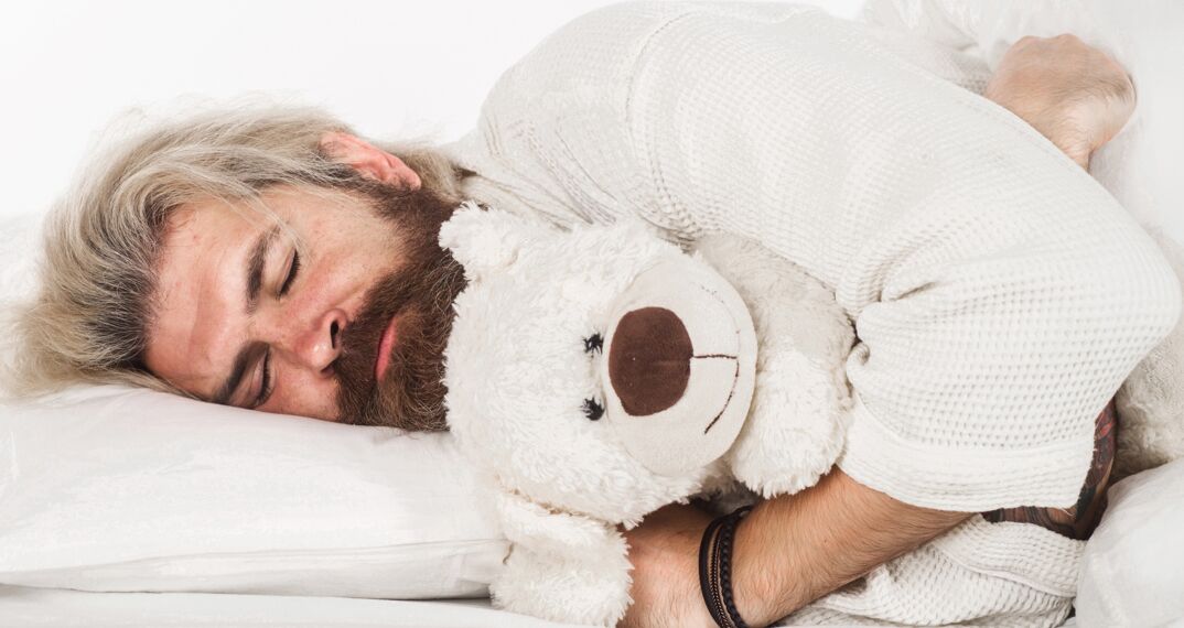 A bearded man sleeping cuddling a stuffed teddy bear. 