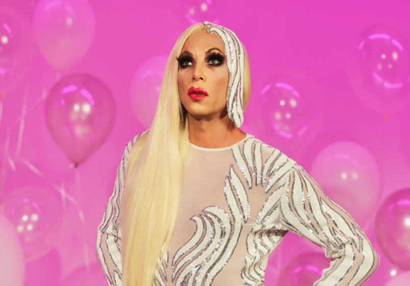 drag queen Sherry Vine wears a white sparkly gown and stands in front of a pink wall and balloons.