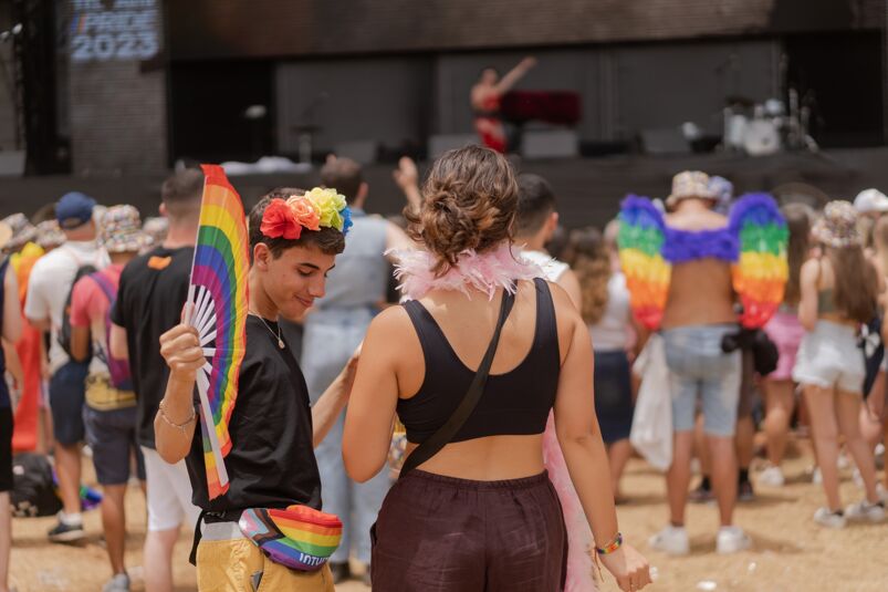 a queer person wearing a rainbow flower crown, fanny pack, and fan at an outdoor Pride festival