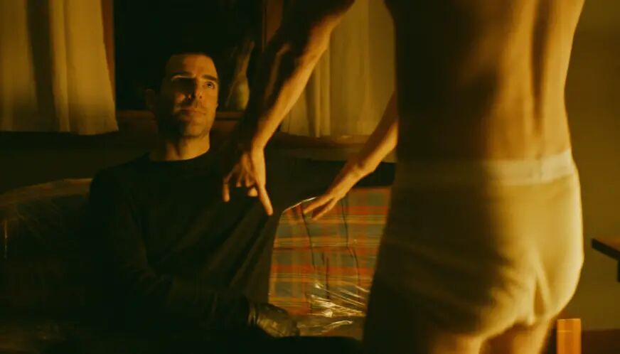 Zachary Quinto sits on a couch and look at a man dancing in white underwear