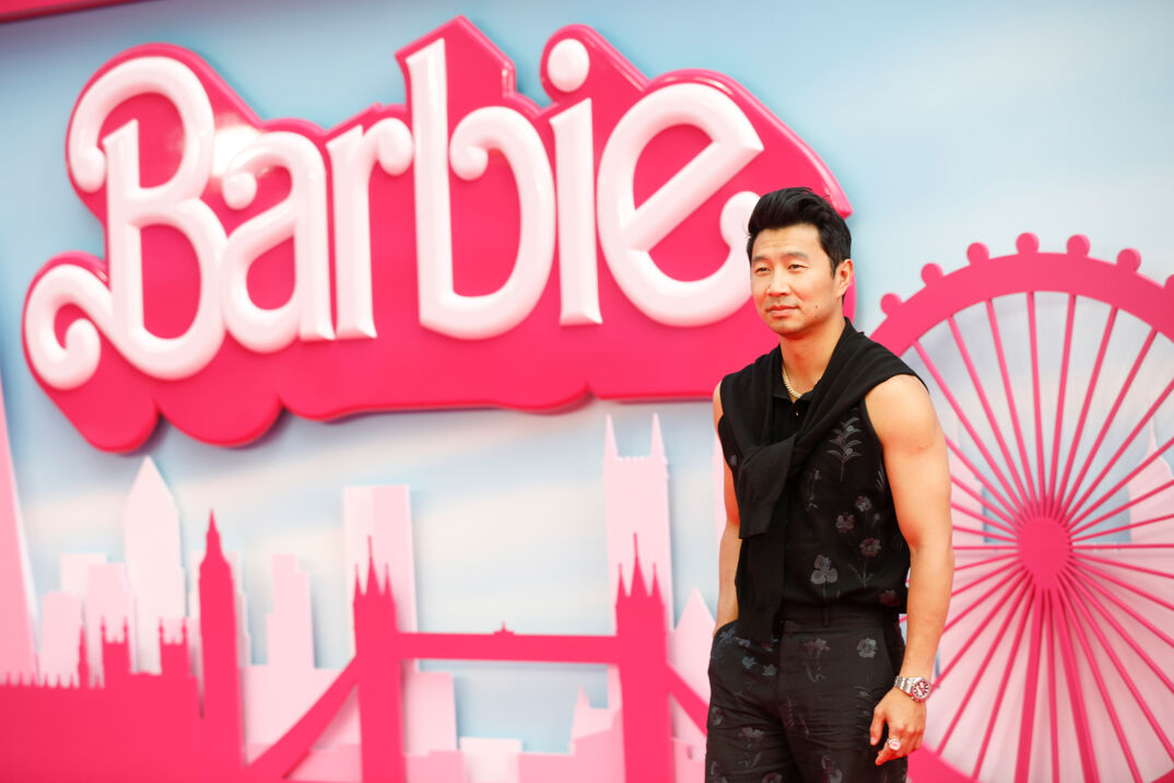 Simu Liu, wearing a black sleeveless jumpsuit and a black sweater tied around his neck, poses in front of the step-and-repeat for 'Barbie' at the European premiere.