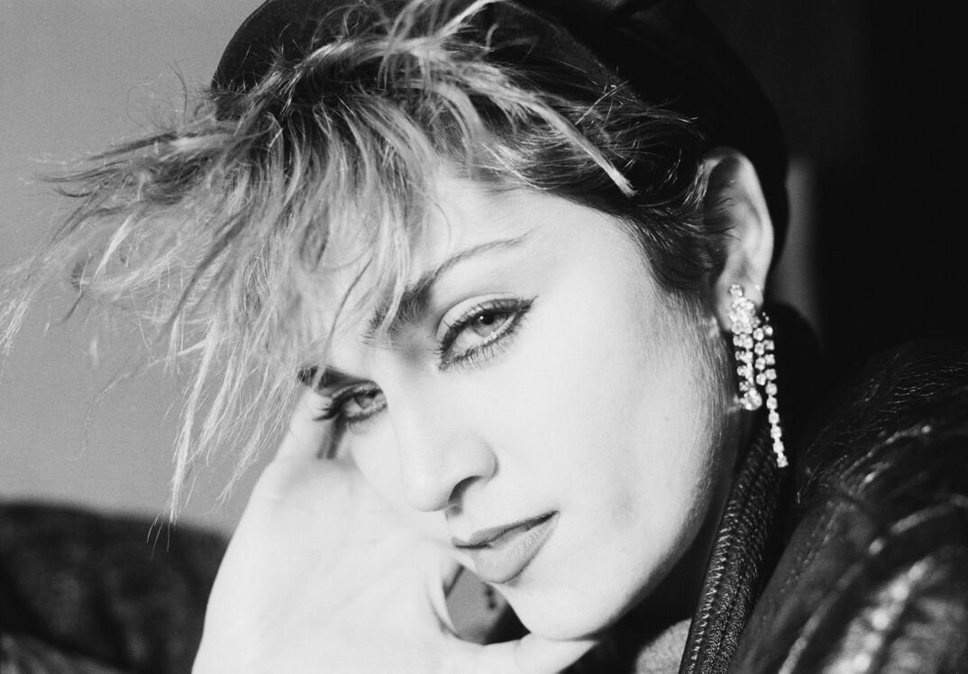 Madonna, wearing a dangling and sparkly earring, looks at the camera seductively in a black and white photo from the '80s, with thick eyeliner and big bangs that stick out in front of her face.