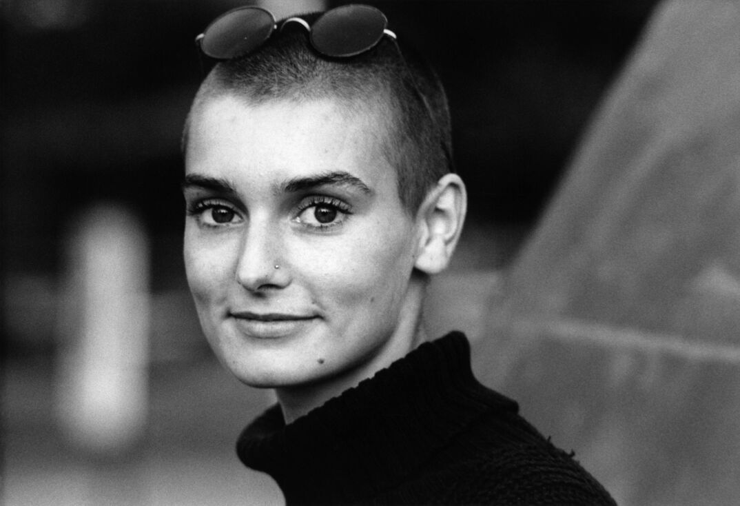 Sinead O'Connor, wearing a nose piercing and sunglasses on her head, softly smiles against a blurry outdoor background in a black-and-white photo.