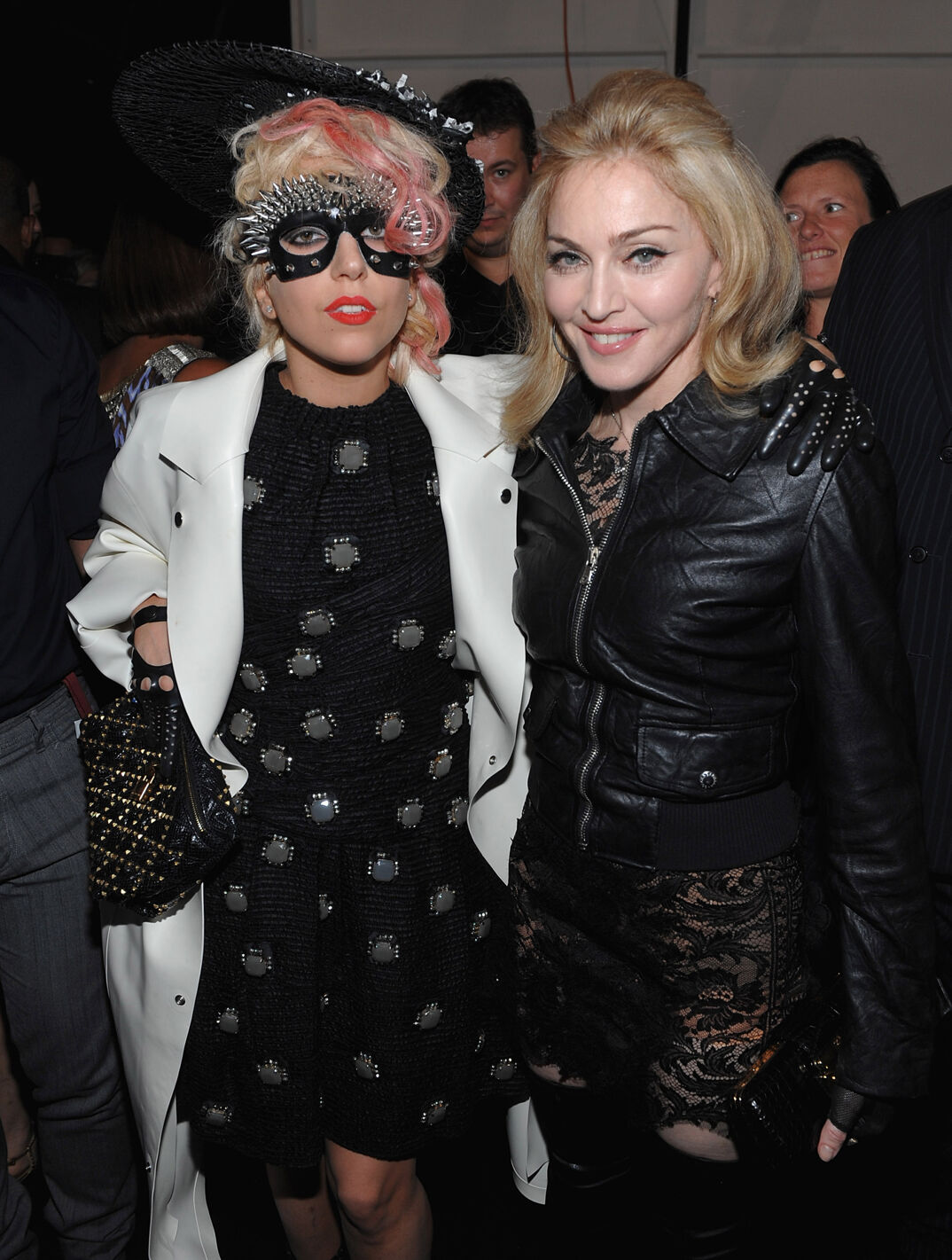 Lady Gaga, with pink-tipped bangs, wears a spiky metal mask around her eyes and leather gloves with her arm around Madonna, who smiles and wears a black leather jacket, at a 2009 fashion event. There's a crowd behind them.