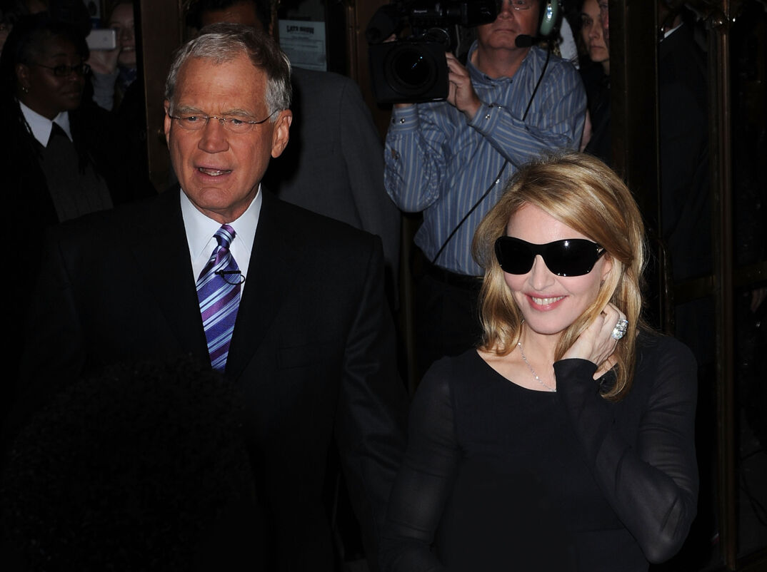 David Letterman, wearing a suit and blue tie, talks to a crowd as he walks alongside Madonna, wearing sun glasses and a tight-fit black shirt on the streets of New York. A cameraman follows them.