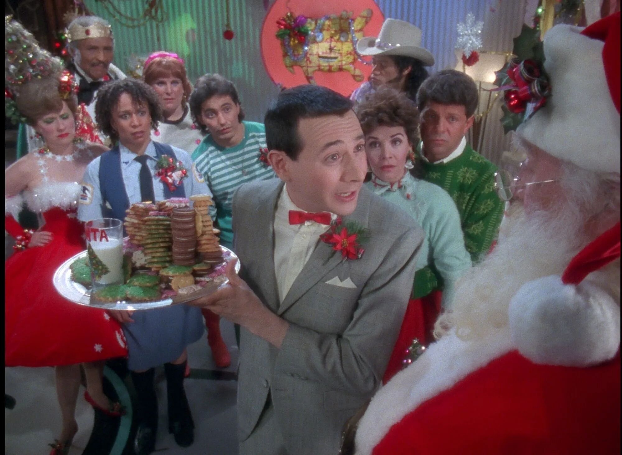 Paul Reubens in Pee Wee’s Playhouse Christmas Special. His character, Pee Wee Herman, stands in front of Santa Claus holding a plate full of cookies and milk. He has a confused look on his face and is surrounded by friends. 