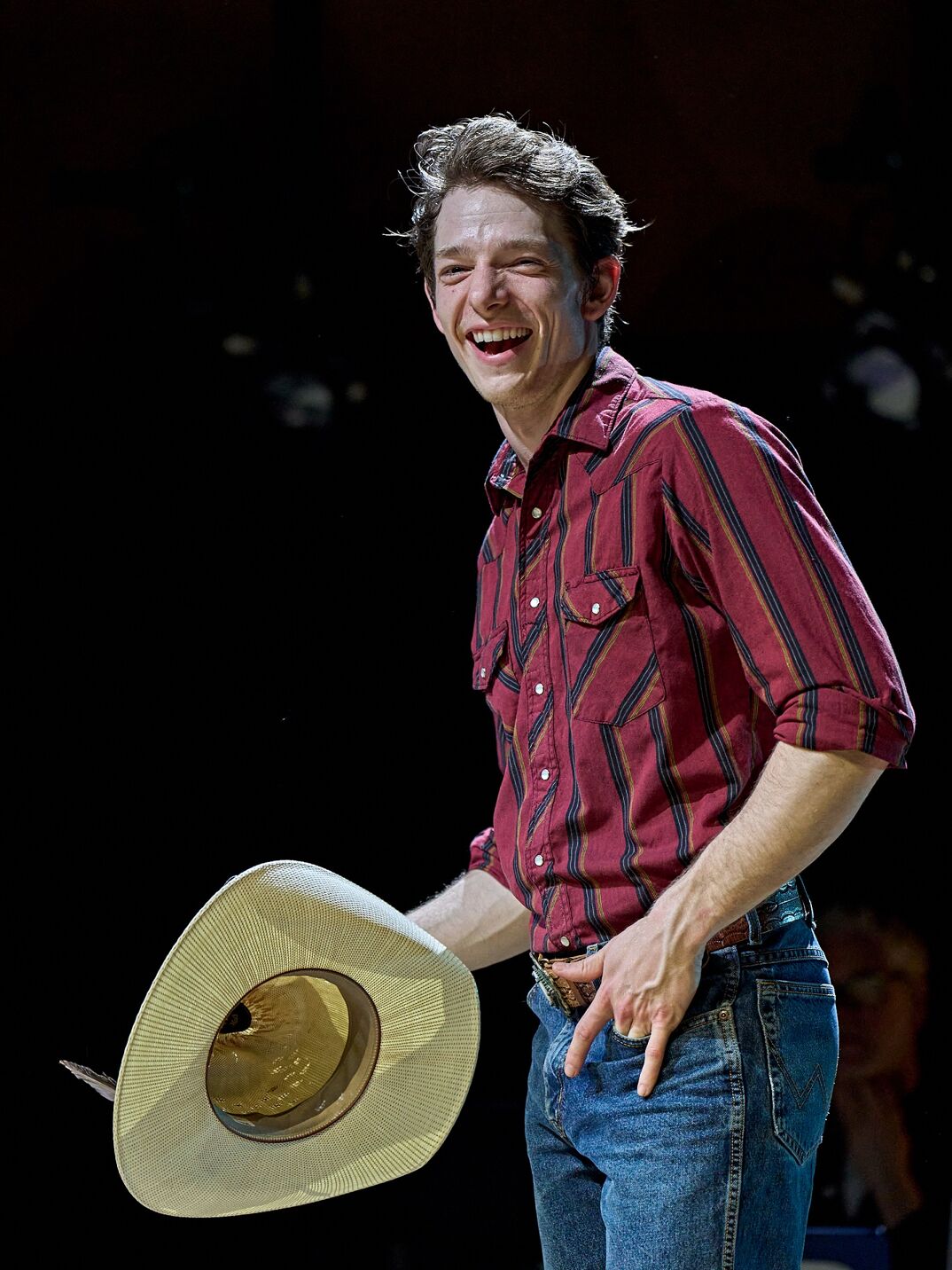 Mike Faist smiles in a red button down shirt and blue jeans, holding a cowboy hat, in a production still from 'Brokeback Mountain.'