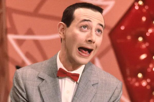 Paul Reubens in character as Pee Wee Herman. His brow is raised in a humorous way and his mouth is open. He is wearing a grey suit jacket with a white undershirt and a red bow tie.