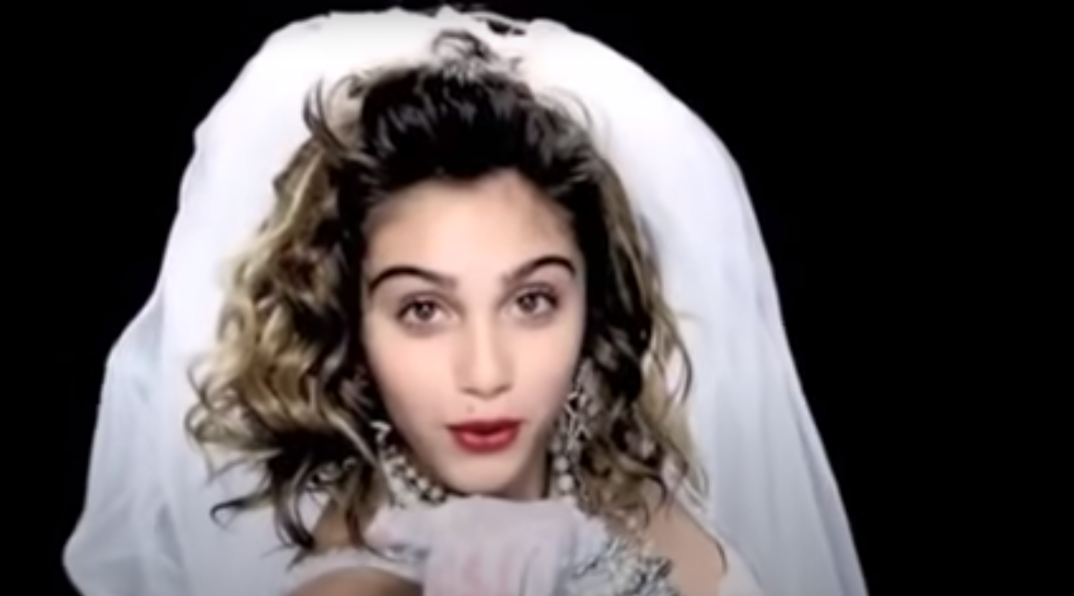 Lourdes Leon, dressed in a white wedding gown with big hair mimicking Madonna's look from "Like a Virgin," blows a kiss towards the camera in a still from Madonna's "Celebration" fan video.