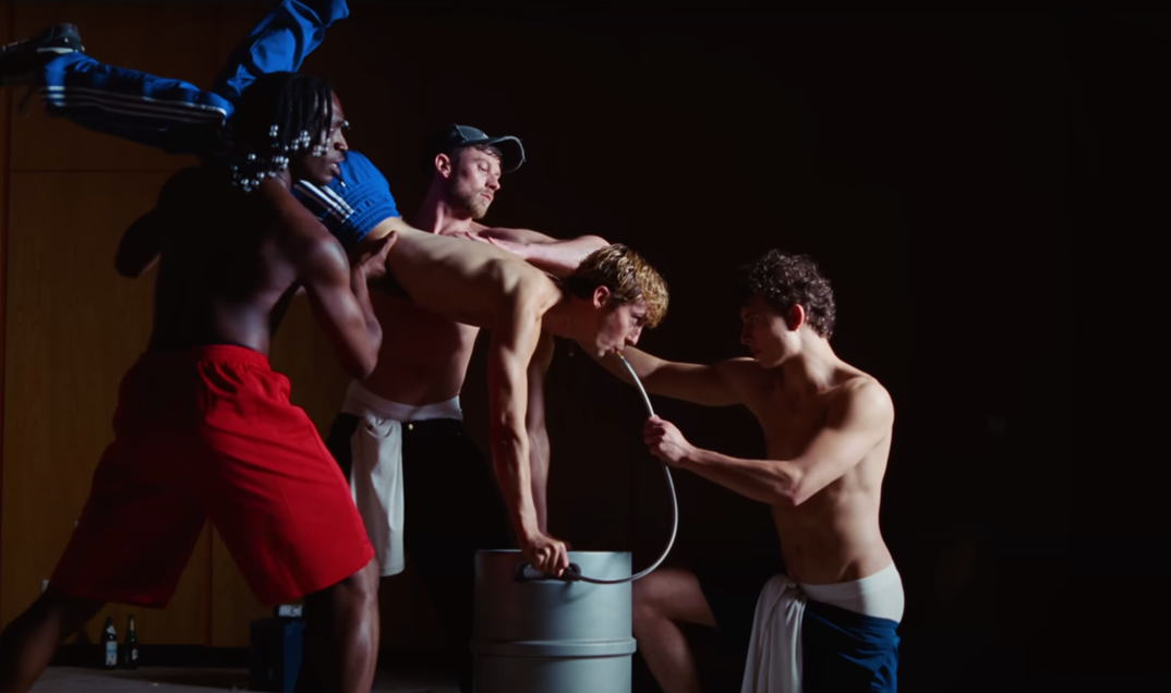 Troye Sivan, shirtless, is hoisted up by a group of shirtless men in a keg stand and theatrically drinks beer from a tube against a black background.