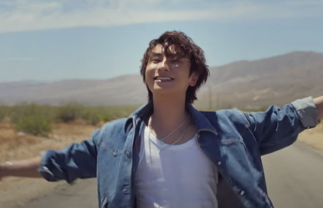 Shinjiro Atae dances with his arms wide open in a white tank top underneath a long sleeve denim shirt. He's smiling and in the desert in this still from his "Into the Light" video.
