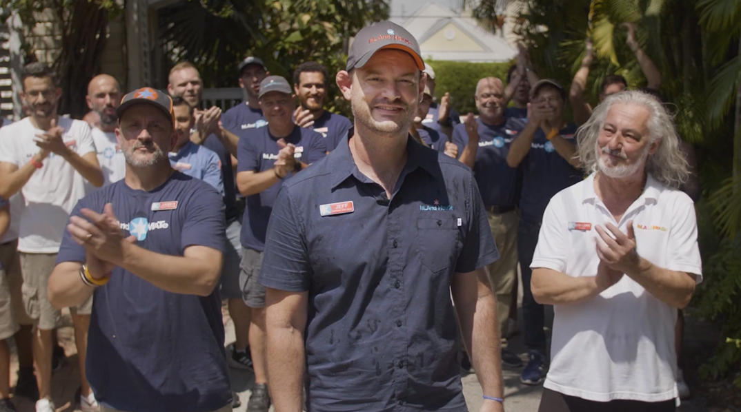 The team of Key West gay resort, Island House, stands in front of the property, featuring beach houses and lush greenery. They mostly wear dark blue shirts with the logo emblazoned on it, and stand applauding.