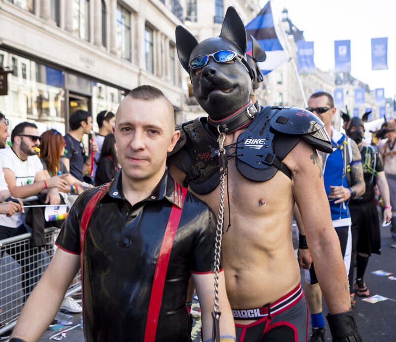 a shirtless man with a pup mask being led by a man with leather polo holding him with a leash.