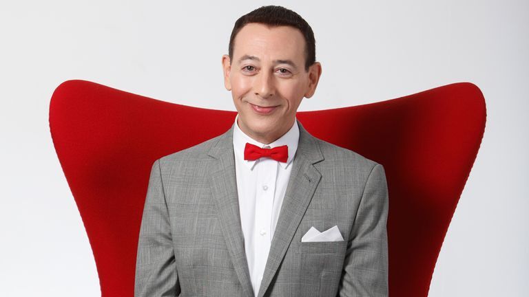Paul Reubens in character as Pee Wee Herman. He is sitting in a red chair against a white background getting his photo taken. He is wearing a grey suit jacket with a white undershirt and a red bow tie. He has a pleasant smile on his face.