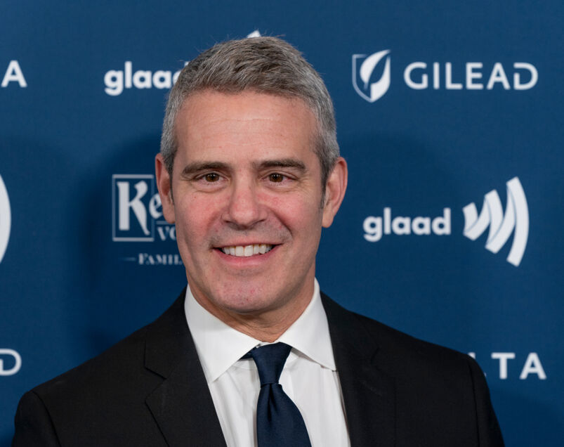 Andy Cohen smiles at the camera at the 30th Annual GLAAD Media Awards in New York. He is wearing a black blazer jacket with a white undershirt and a navy blue tie. His hair is grey, but he is handsome.