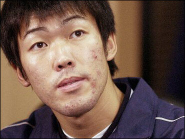 Kazuhito Tadano looks at the camera while being interviewed with CBS News. He is wearing a blue track jacket and a white undershirt. He has blemishes on his face and black hair.