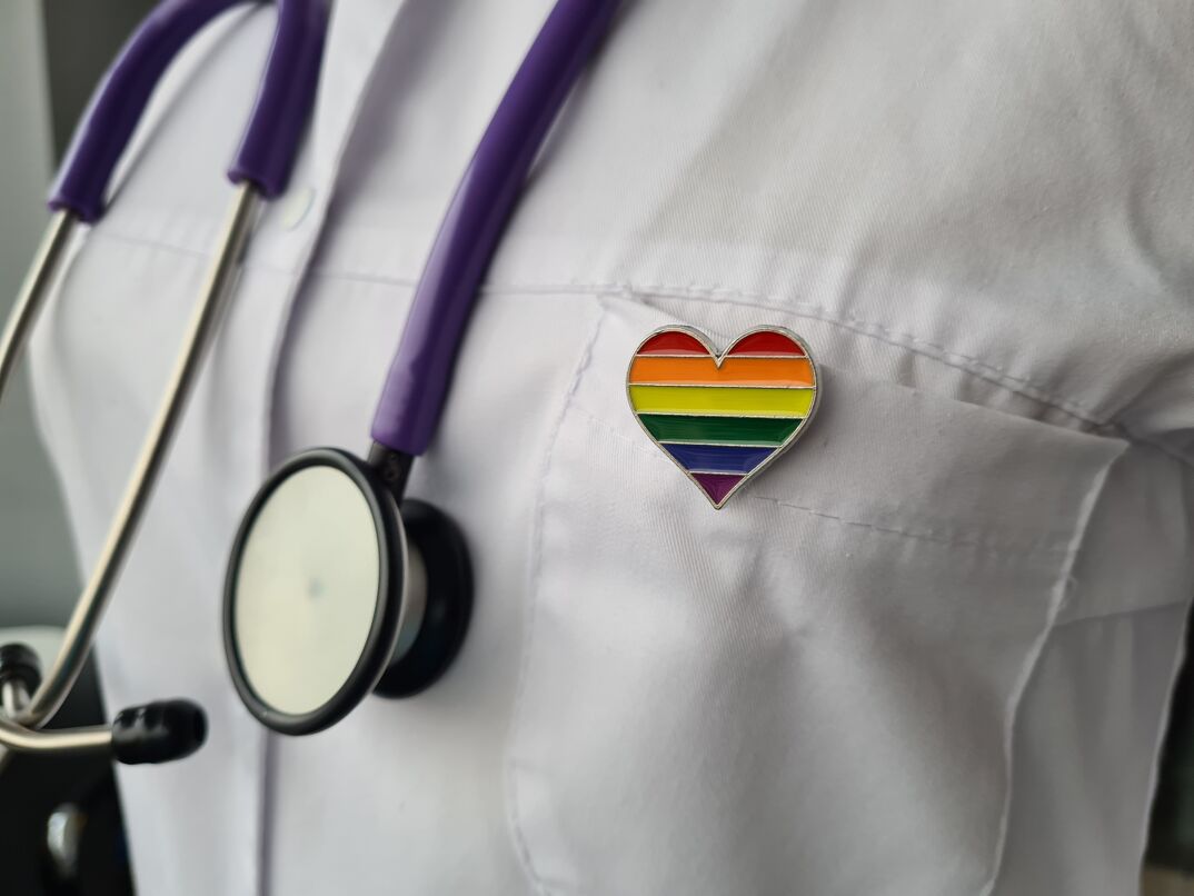 close-up of a person wearing a white doctor's lab coat with a stethoscope and a rainbow heart pin to represent that they are an inclusive healthcare provider for LGBTQ+ people
