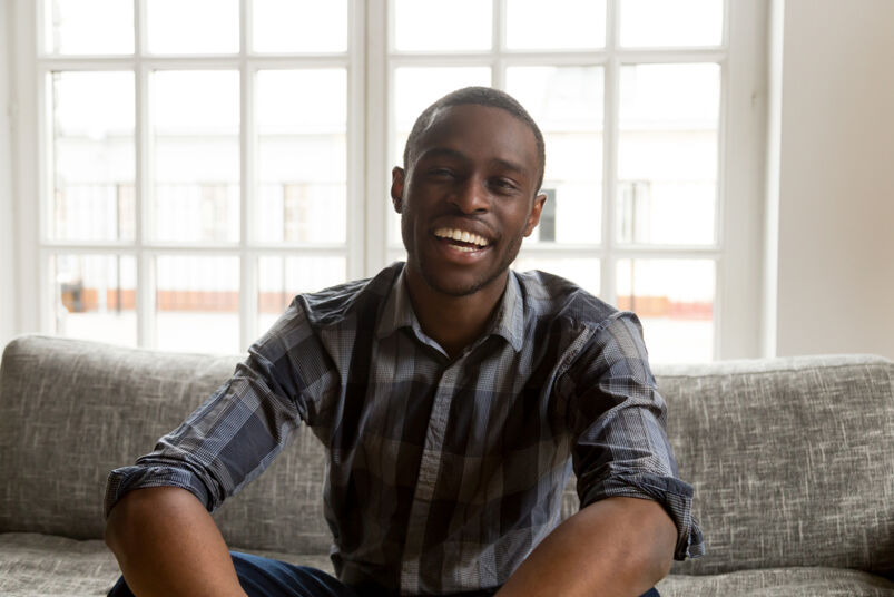 Facts about semen: Smiling Black male wearing a dark colored striped shirt looking at camera sitting on couch.