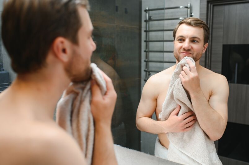 Shirtless Caucasian man with a towel around his torso looking at himself in mirror admiring his smooth skin.