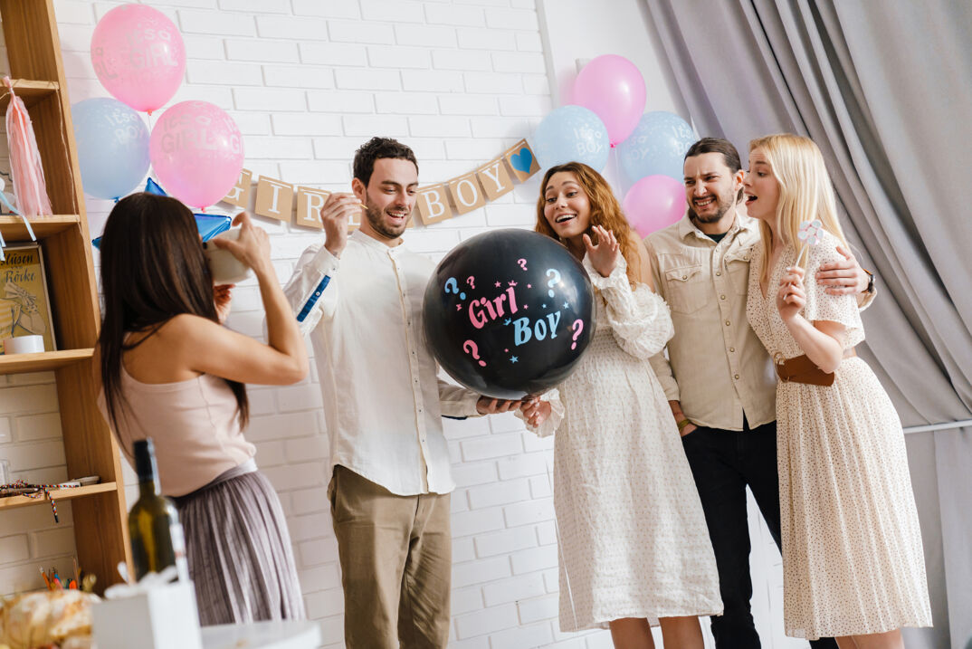 a group of people at a gender reveal party standing around a black balloon with the words girl in pink and boy in blue written on it ready to learn if their child will be AMAB or AFAB