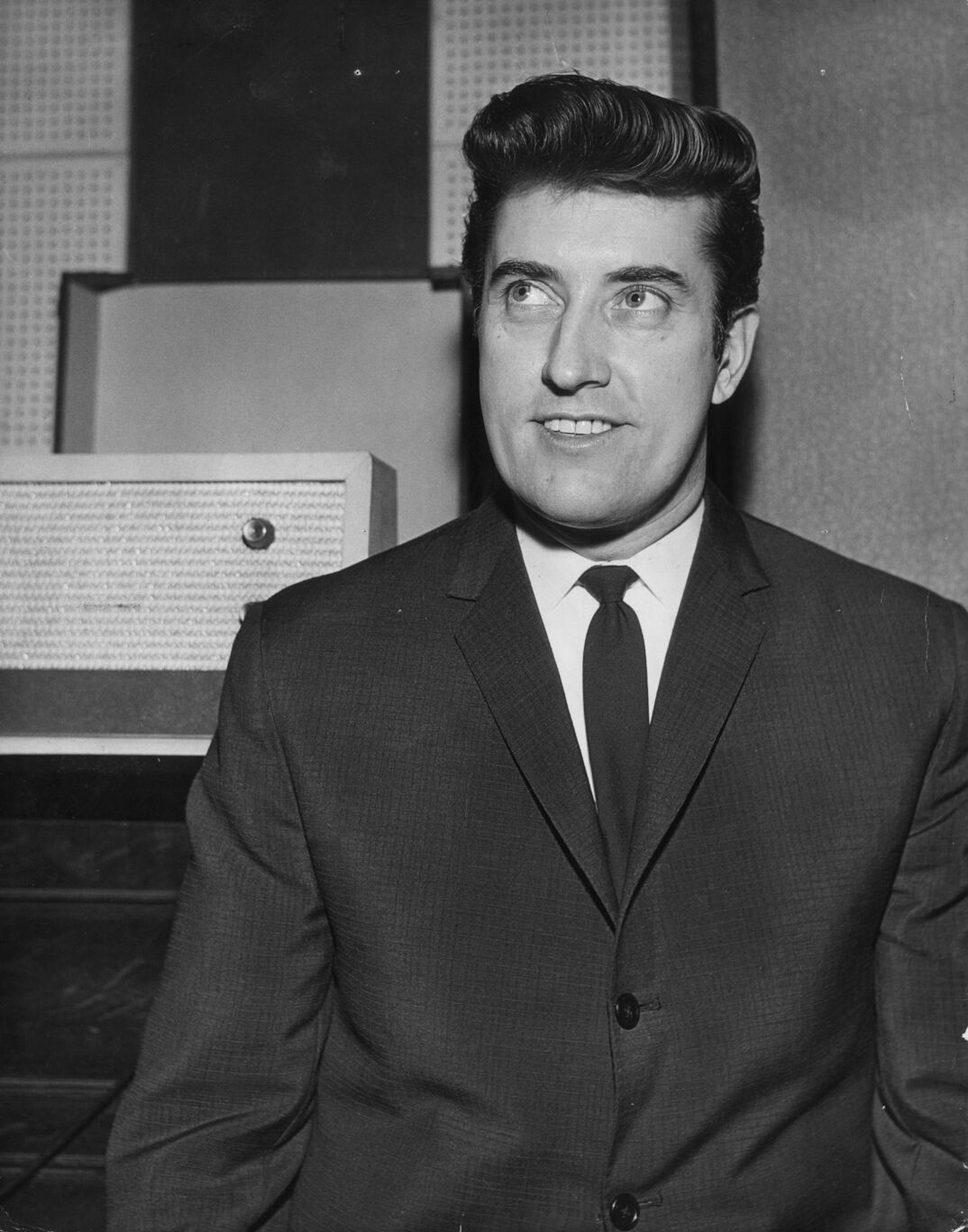 Black-and-white photo of producer Joe Meek, pictured in a recording studio in 1963. He has perfectly coiffed hair and smiles while looking up. He wears a black suit, buttoned up and a white dress shirt with a black tie.