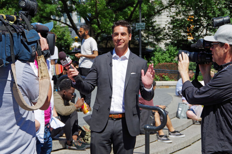 Jesse Watters standing outride in a dark suit jacket and white dress shirt holding a Fox News mic in front of a camera.