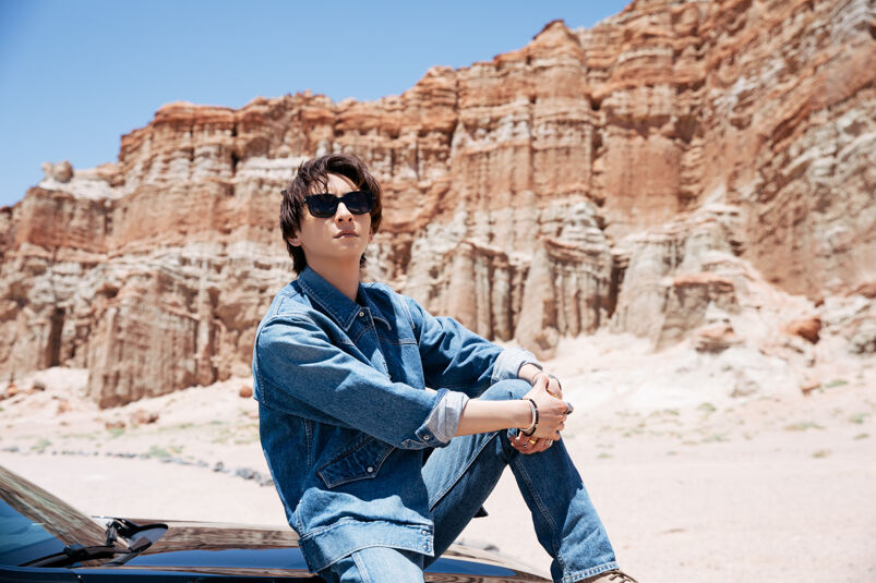 Shinjiro Atae sits on top of a black car in the middle of a scenic desert. He wears a denim long sleeve shirt and jeans and sun glasses. He looks into the camera thoughtfully.