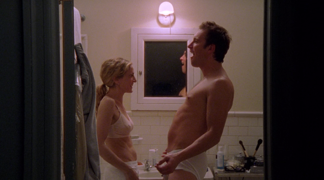 Carrie Bradshaw (Sarah Jessica Parker) stands across from Aidan Shaw  (John Corbett) in front of their bathroom mirror. The two are laughing and brushing their teeth, both wearing white matching briefs.
