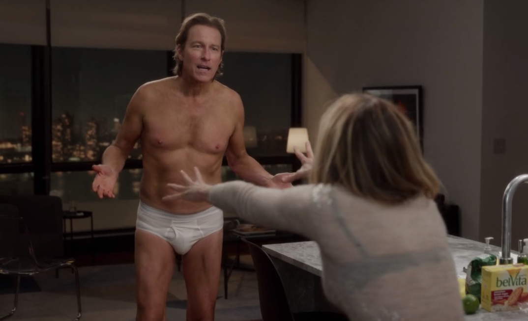 Aidan Shaw (John Corbett) stands shirtless in white briefs talking to Carrie Bradshaw (Sarah Jessica Parker) in an apartment kitchen in an episode of 'And Just Like That.' The two are in a confused conversation.
