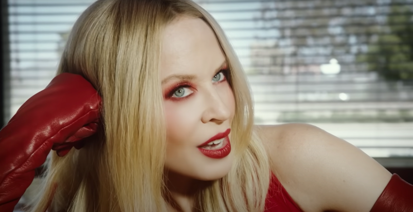 Kylie Minogue, wearing bright red lipstick and eye shadow, leans her head against a red glove as she sits at the bar at a diner in front of a window in the "Padam Padam" music video.