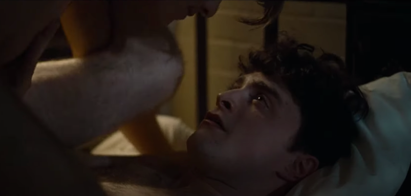 Daniel Radcliffe looks up at a man, whose face in partially out of frame, as he lays on his bed with his legs spread over the man's shoulders in a darkened bedroom in a scene from 'Kill Your Darlings.'