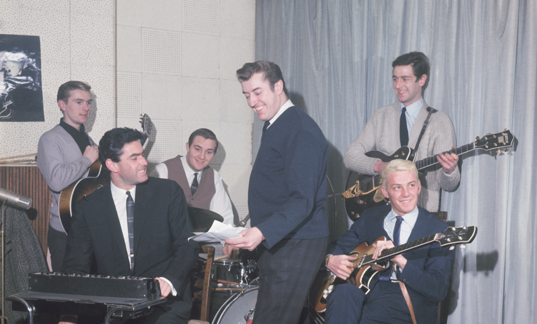 British instrumental pop  group including guitarists Alan Caddy and George Bellamy, drummer Clem Cattini, keyboardist Roger Lavern and bassist Heinz Burt sit playing their instruments in 1963 at producer and manager Joe Meek's house. They smile in different directions.