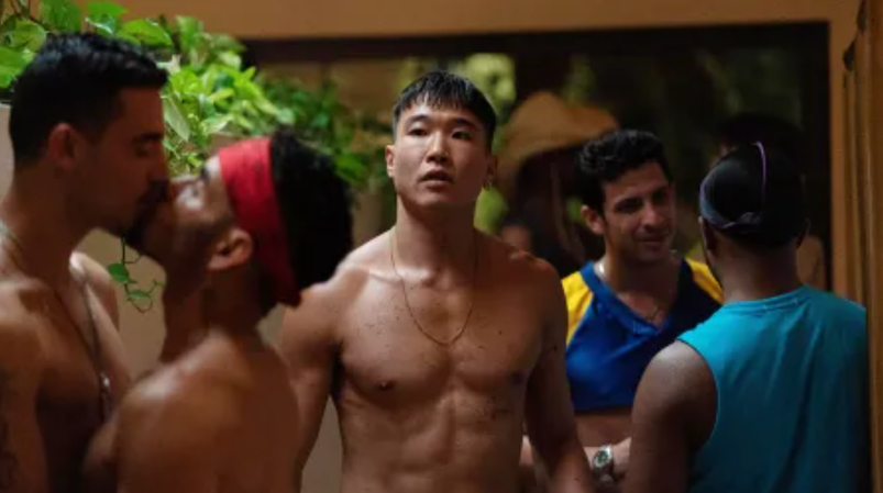 In a still from 'Fire Island,' Joel Kim Booster, shirtless and wearing a chain necklace, walks through a gay party. There's a lush greenery behind him, and a shirtless couple making out on his left. Behind him, another couple flirts. 