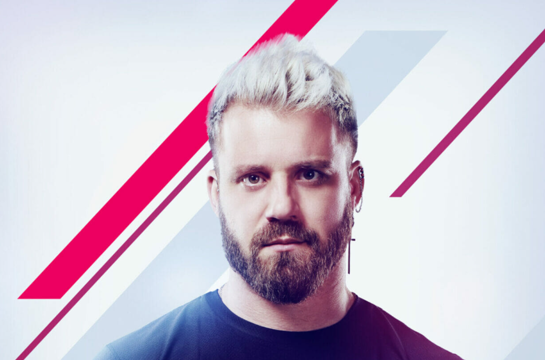 Paulie Calafiore, with spiky bleached hair, poses in front of a white and red background. He wears a black tee, has a dark brown beard, and wears a long cross earring on his right ear. 