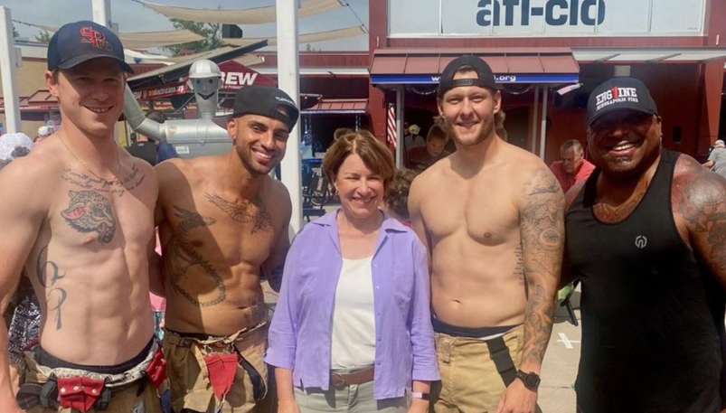 Outside at the Minnesota State Fair, Senator Amy Klobuchar stands wearing a purple shirt over a white tank top, khaki pants, and modest shoes. She smiles as she's surrounded by muscular shirtless firefighters, stripped down to just the pants of their turnout gear. They are smiling and stand with their arms around her back.