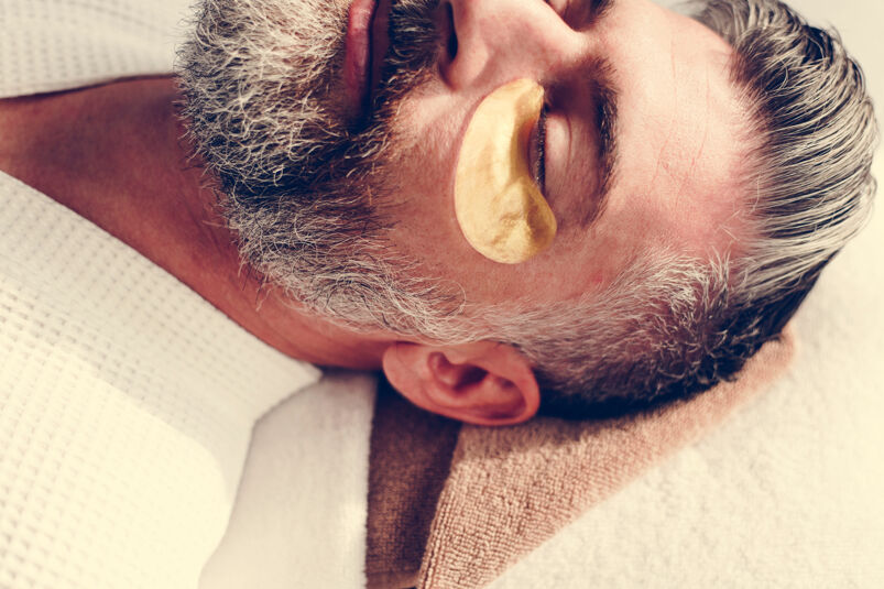 An older man with grey hair and a beard is doing an undereye hydration mask. 