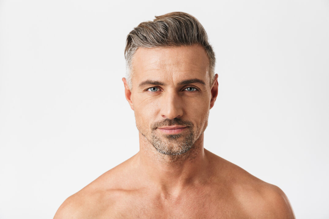A man with blue eyes and grey hair stares at the camera shirtless. 